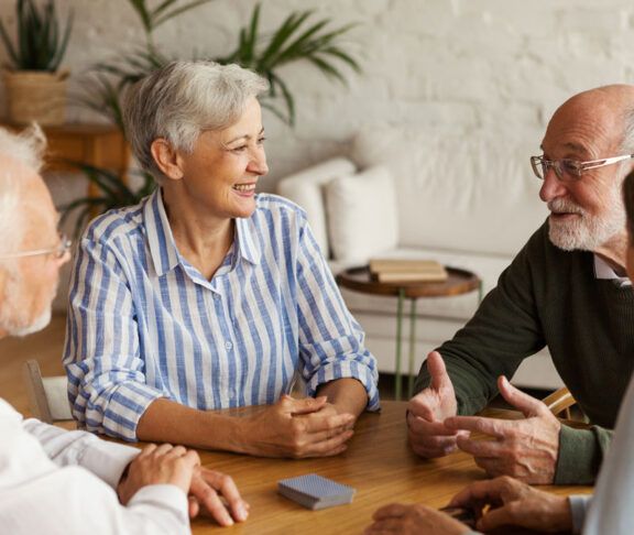 Group of four cheerful senior friends, two men and two women, sitting at table and enjoying talk after playing cards in assisted living home