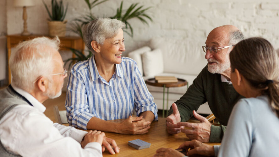 Group of four cheerful senior friends, two men and two women, sitting at table and enjoying talk after playing cards in assisted living home
