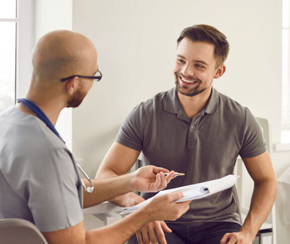 Portrait of a happy smiling young man patient in medical office listening to doctor holding report file with appointment and giving consultation during medical examination in clinic