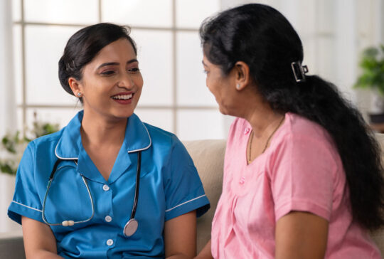 Side view shot of happy indian nurse with talking with recovered woman patient on sofa during home visit - concept of health care support, consultation and professional occupation