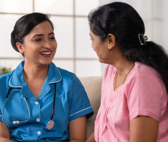 Side view shot of happy indian nurse with talking with recovered woman patient on sofa during home visit - concept of health care support, consultation and professional occupation