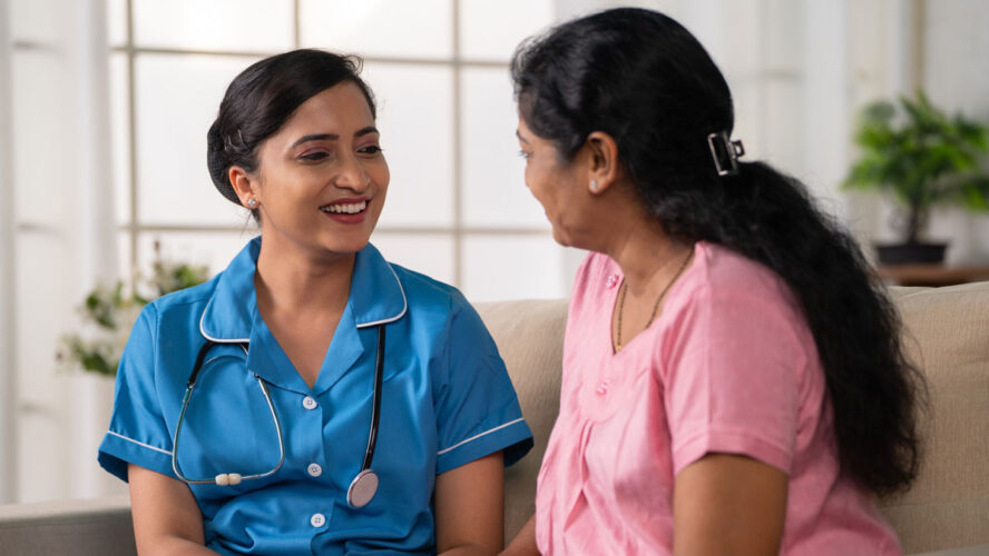 Side view shot of happy indian nurse with talking with recovered woman patient on sofa during home visit - concept of health care support, consultation and professional occupation