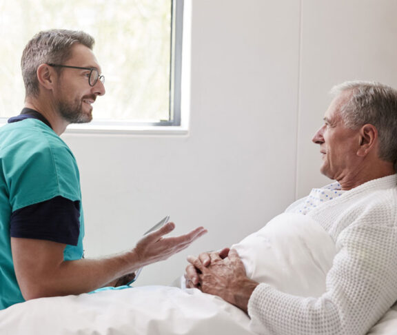 Surgeon Visiting And Talking With Senior Male Patient In Hospital Bed In Geriatric Unit
