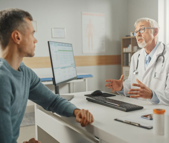Middle Aged Family Doctor is Talking with Young Male Patient During Consultation in a Health Clinic. Senior Physician in Lab Coat Sitting Behind a Computer Desk in Hospital Office