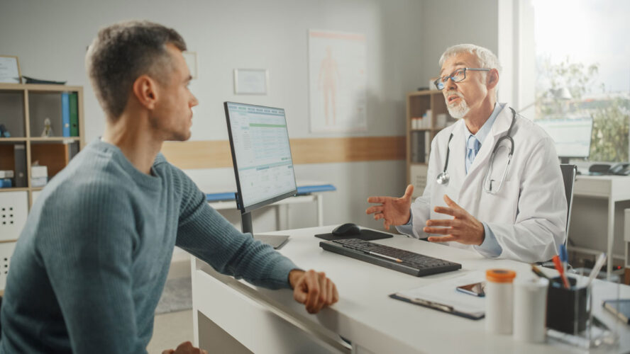 Middle Aged Family Doctor is Talking with Young Male Patient During Consultation in a Health Clinic. Senior Physician in Lab Coat Sitting Behind a Computer Desk in Hospital Office