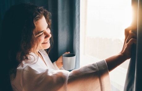 Curly haired lady in white bathrobe opens curtains to look outside large window drinking hot coffee in dark hotel room at sunrise time