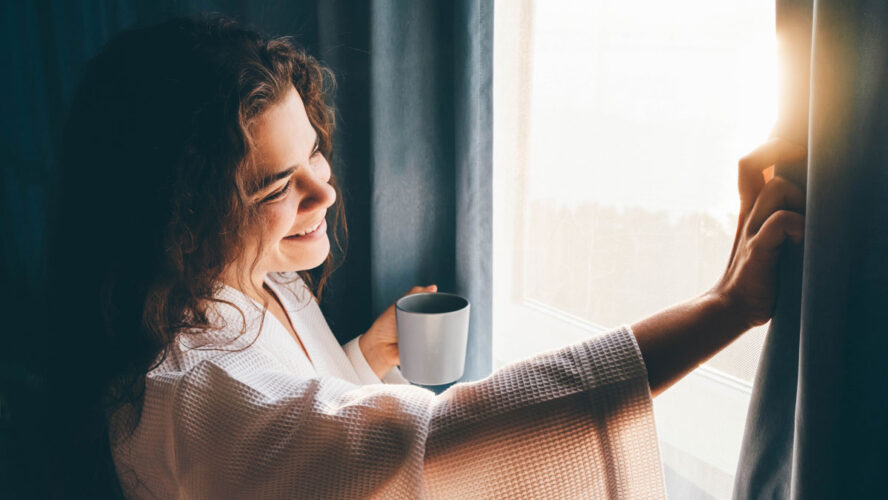 Curly haired lady in white bathrobe opens curtains to look outside large window drinking hot coffee in dark hotel room at sunrise time
