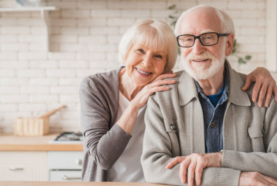 Portrait of caucasian smiling senior old elderly couple family spouses grandparents looking at camera, embracing hugging with love and care at home kitchen