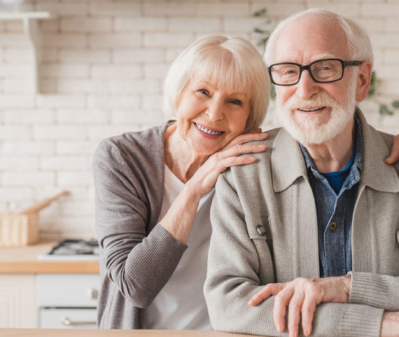 Portrait of caucasian smiling senior old elderly couple family spouses grandparents looking at camera, embracing hugging with love and care at home kitchen