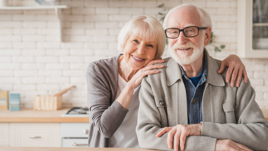 Portrait of caucasian smiling senior old elderly couple family spouses grandparents looking at camera, embracing hugging with love and care at home kitchen