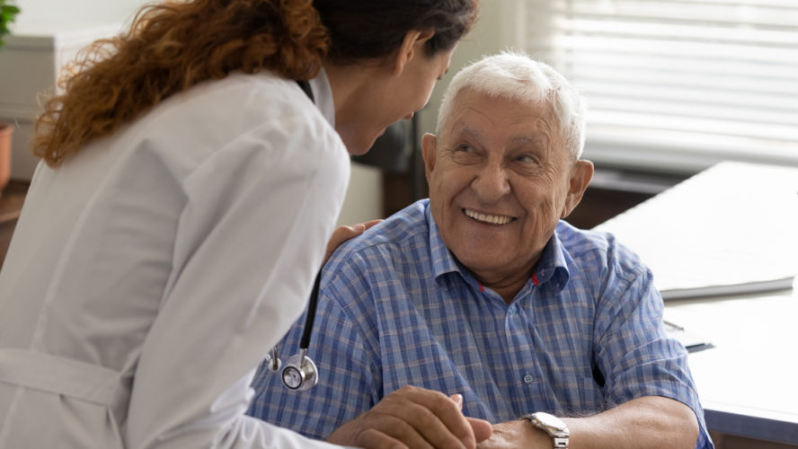 Friendly young female doctor and optimistic elder 80s man meeting in office for consultation. GP therapist holding hands of old patient, giving sympathy, support, advice about treatment