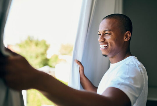 Shot a handsome young man opening his bedroom curtains in the morning