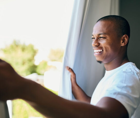 Shot a handsome young man opening his bedroom curtains in the morning