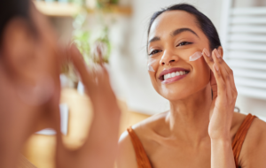 Smiling mixed race young woman applying moisturizer on her face in bathroom