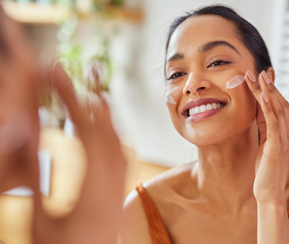 Smiling mixed race young woman applying moisturizer on her face in bathroom