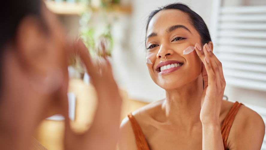 Smiling mixed race young woman applying moisturizer on her face in bathroom